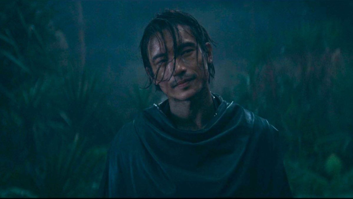 Manny Jacinto as Qimir in a scene from the Disney+ Star Wars show, 'The Acolyte.' He is a Filipino-Chinese man with chin-length dark hair hanging in his face as he stands in the rain dressed all in black and looking smug.
