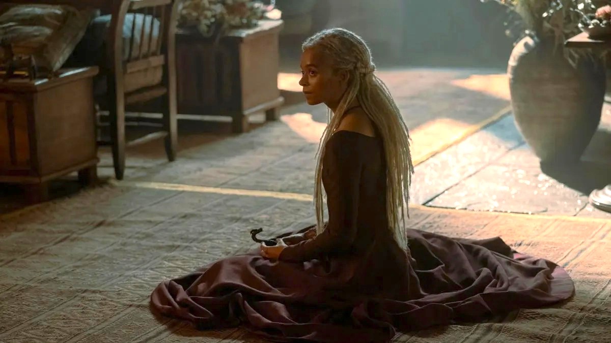 Phoebe Campbell as Rhaena Targaryen sits on the floor clutching a wooden toy dragon in her hands in House of the Dragon season 2