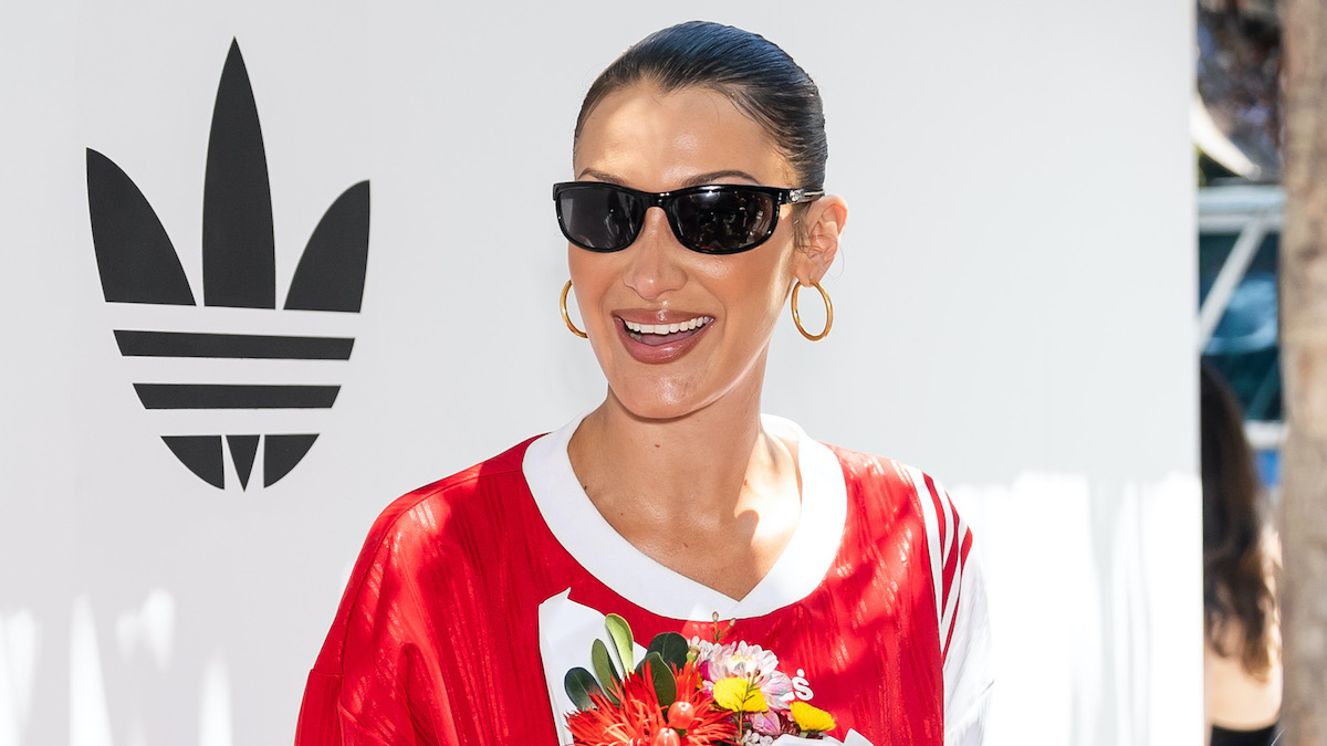 Bella Hadid grins, wearing sunglasses next to an event backdrop wth the Adidas logo on it.