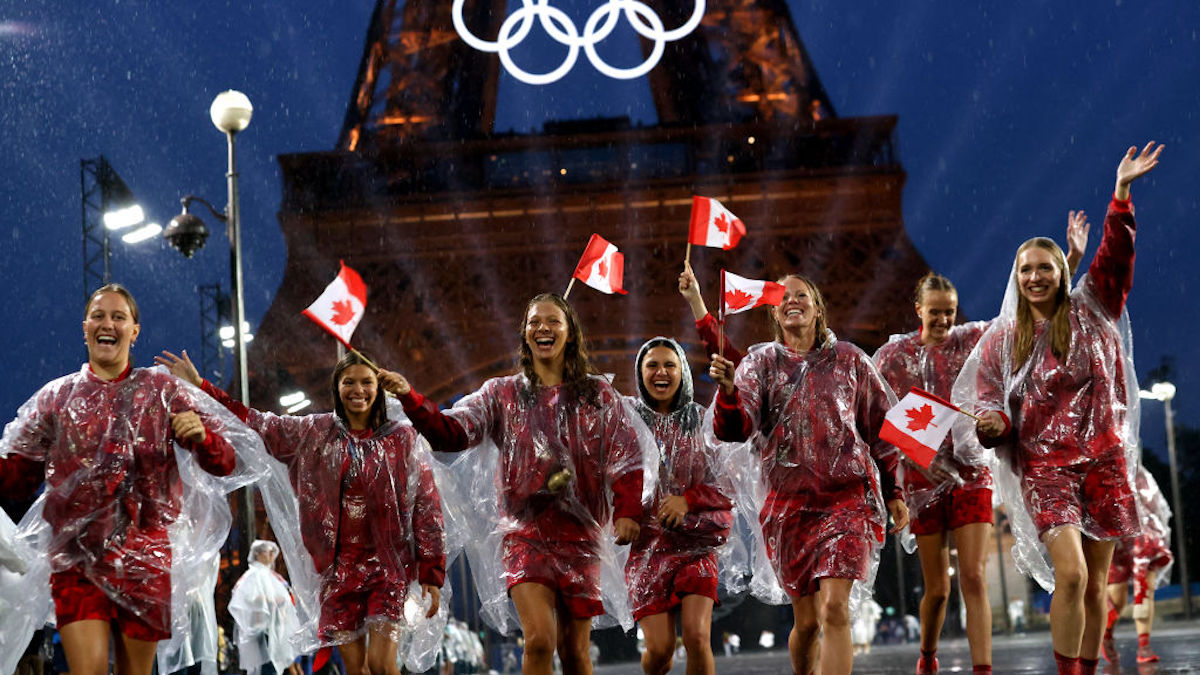 A group of female athletes holding small Canadian flags run and grin in front of the eiffel tower