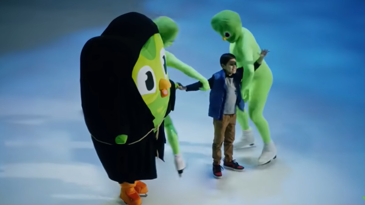 The Duolingo owl in a black robe and ice skates, standing on the ice, oversees two people in green body suits, ice skates, and Duo masks, carrying a child away