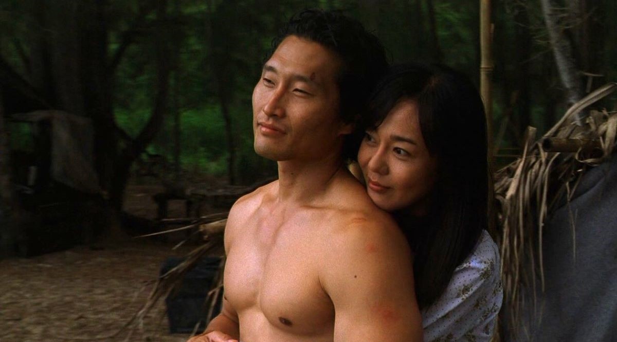 Image of Daniel Dae Kim as Jin and Yunjin Kim as Sun in a scene from ABC's 'Lost.' Jin is a Korean man with shaggy dark hair standing shirtless. Sun is a Korean woman with long dark hair and bangs. She's wearing a white, long-sleeved blouse as she stands behind him with her arms wrapped around him and her head on his shoulder. They are both looking out at something and smiling.