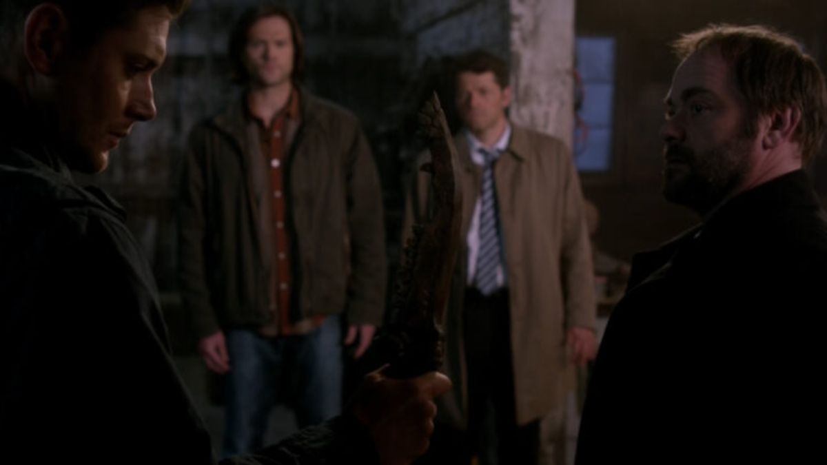 Jensen Ackles as Dean Winchester holds the First Blade against Mark Sheppard's Crowley as Misha Collins' Castiel and Jared Padalecki's Sam Winchester look on