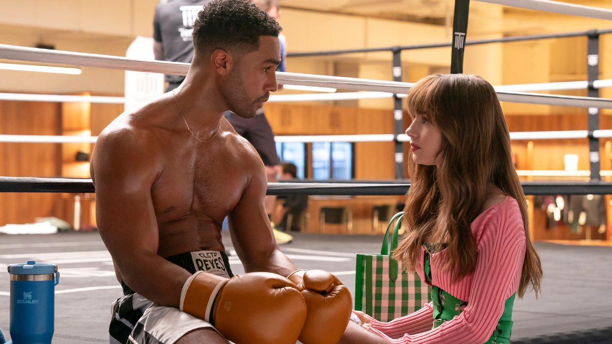 Lucien Laviscount as Alfie sits in the boxing ring with gloves on as Lily Collins as Emily talks to him in Emily in Paris season 4