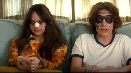 a girl with a bandage on her nose sits on the couch next to her brother, whose face has been drawn on