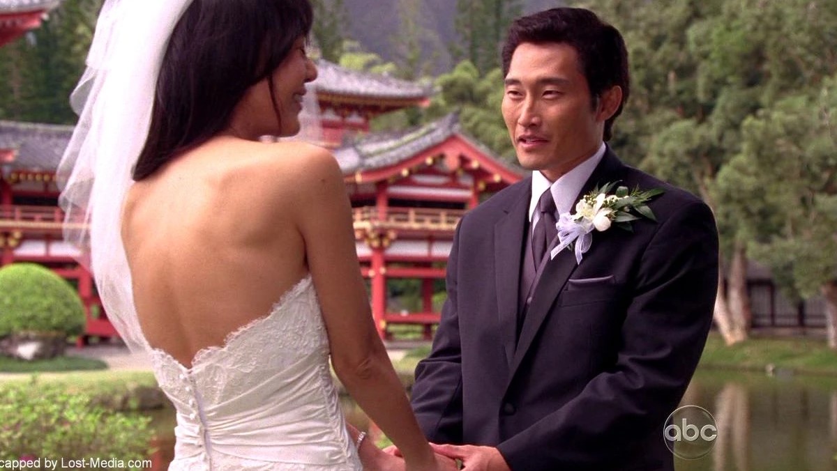 Image of Yunjin Kim as Sun and Daniel Dae Kim as Jin in a scene from ABC's 'Lost.' Sun is a Korean woman with long, dark hair, and we see her from behind wearing a wedding dress and veil with her face turned smiling to the side. Jin is a Korean man with short, dark hair wearing a dark suit and a boutonniere with a white flower in his lapel as he holds Sun's hands. They are standing oudoors at the temple where they're getting married.