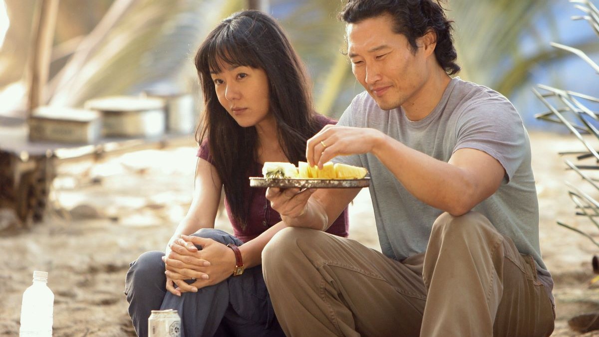 Image of Yunjin Kim as Sun and Daniel Dae Kim as Jin in a scene from ABC's 'Lost.' Sun is a Korean woman with long dark hair and bangs wearing a maroon cap-sleeved shirt and grey pants. Jin is a Korean man with shaggy dark hair wearing a grey t-shirt and khakis. They are sitting at their beach camp. Jin is eating mango off a tray as Sun looks out at something.