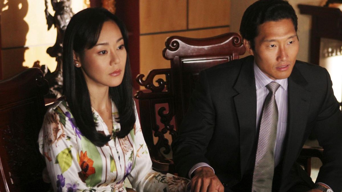 Image of Yunjin Kim as Sun and Daniel Dae Kim as Jin in a scene from ABC's 'Lost.' Sun is a Korean woman with long, dark hair, wearing a white, floral print silk blouse.. Jin is a Korean man with short, dark hair wearing a dark suit and a grey buttondown with white stripes and a grey tie. They are sitting in an office holding hands as they look at someone speaking to them.