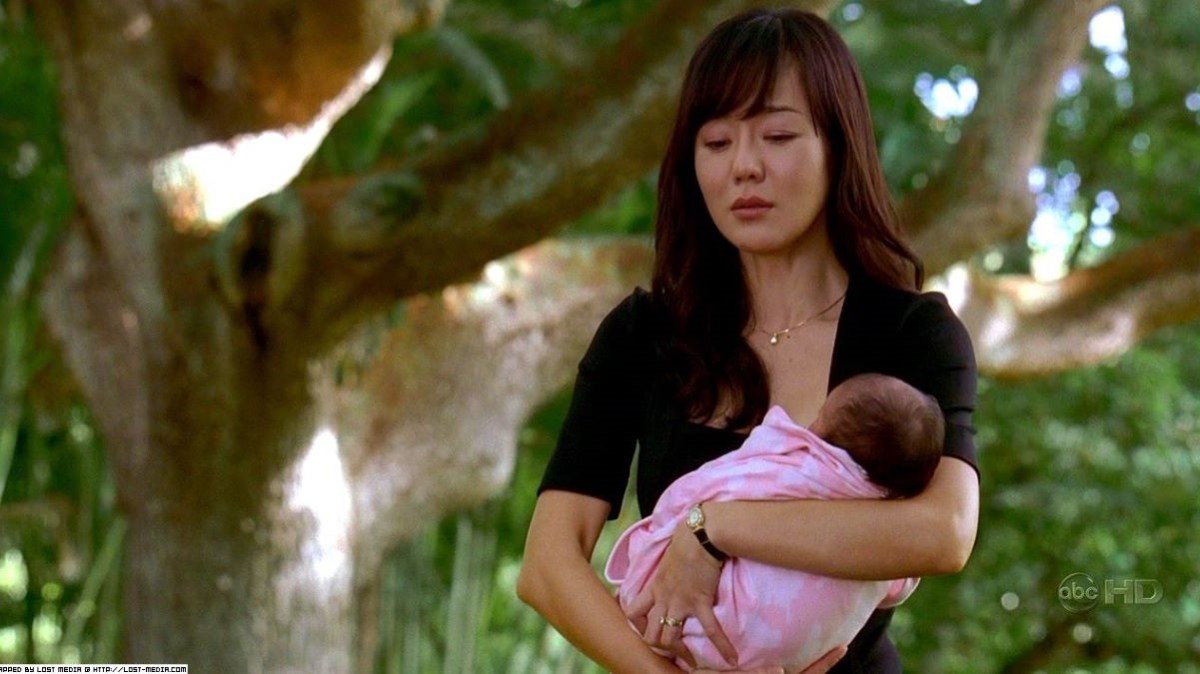 Image of Yunjin Kim as Sun in a scene from ABC's 'Lost.' Sun is a Korean woman with long dark hair and bangs wearing a black dress and holding a newborn baby in a pink blanket. She is outside near some trees and is about to cry.