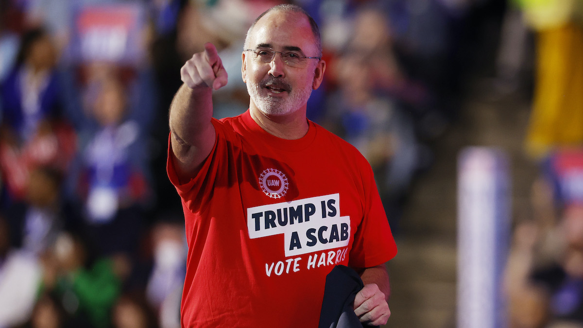 United Auto Workers President Shawn Fain speaks onstage at the DNC wearing a red t-shirt reading "Trump is a scab. Vote Harris"