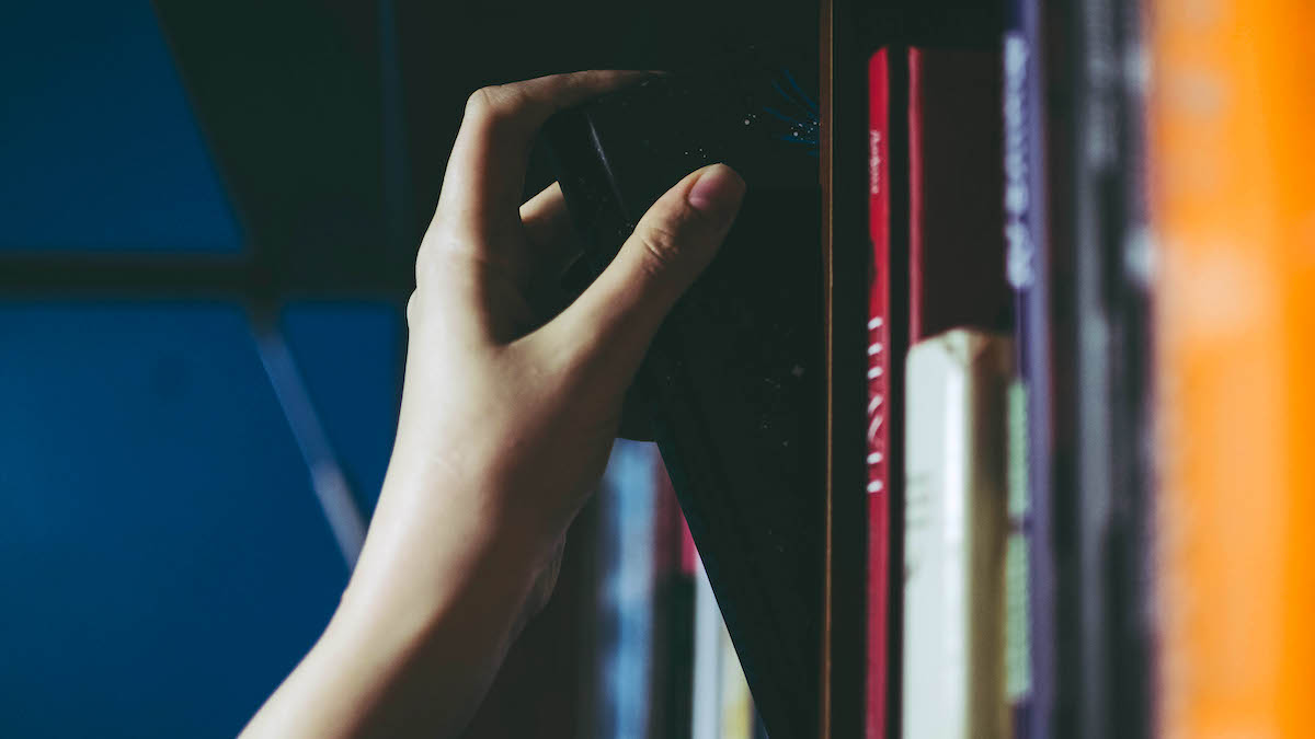 Pale white hand of a girl picking a black book on a shelf.