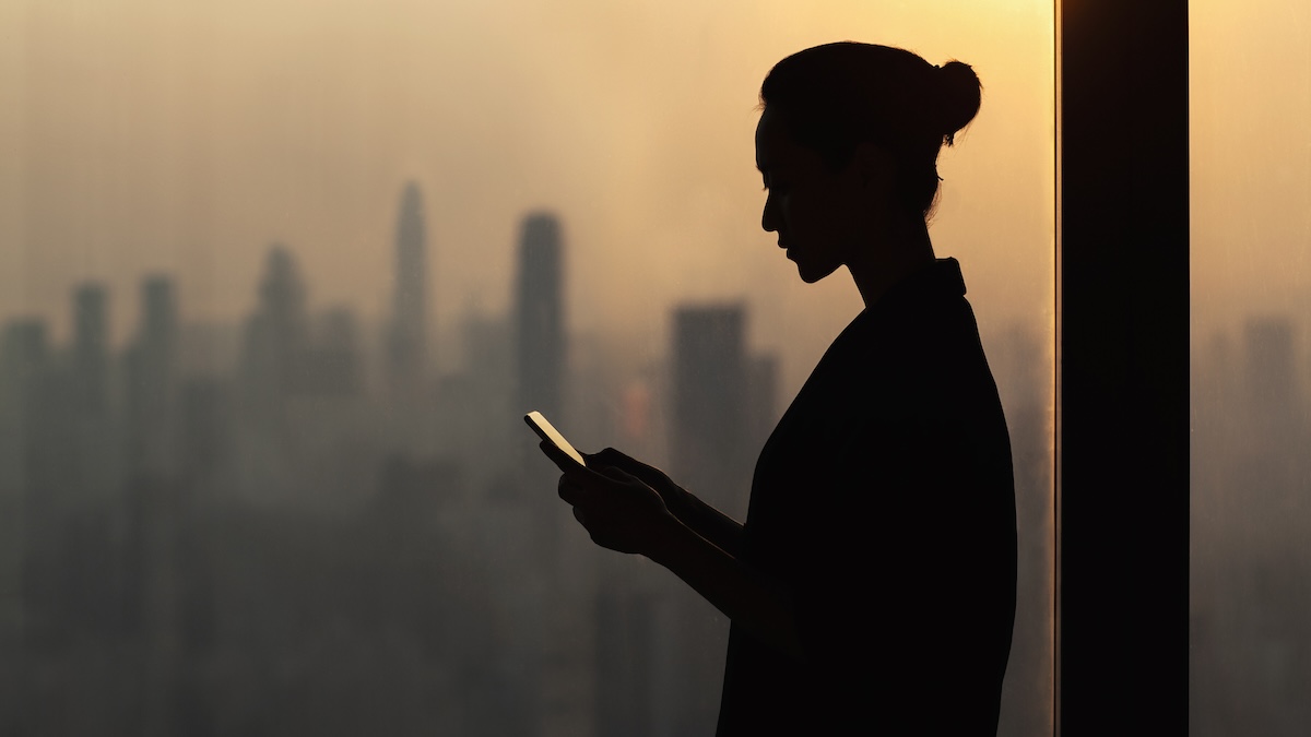 Silhouette of young woman using smartphone next to window with cityscape