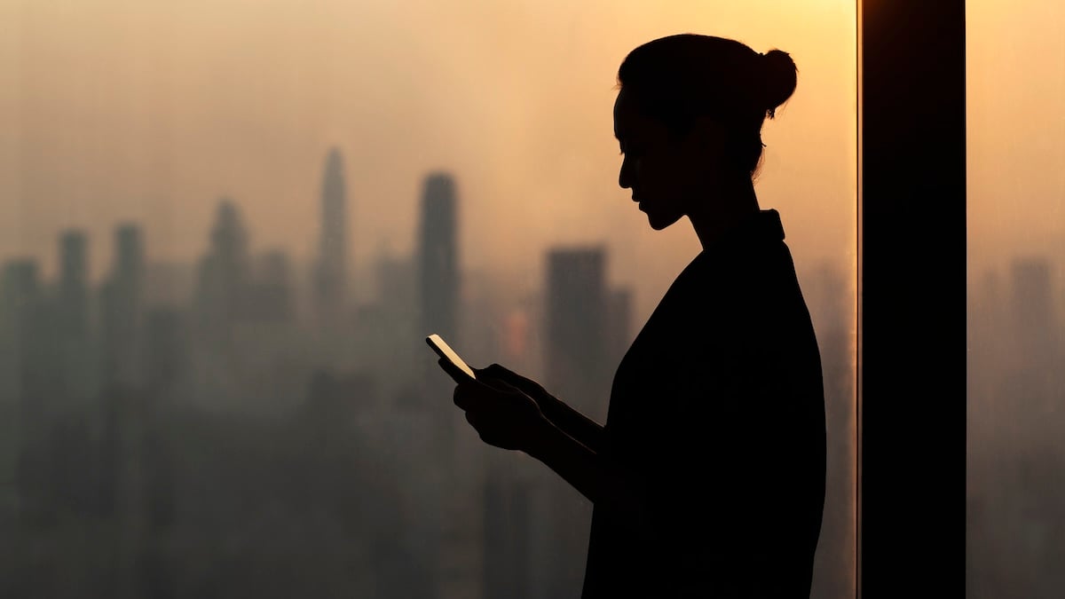 Silhouette of young woman using smartphone next to window with cityscape