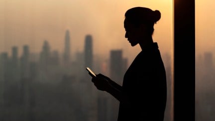 Silhouette of young woman using smartphone next to window with cityscape