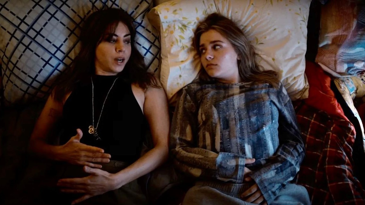 Aubrey Plaza as Older Elliott and Maisy Stella as Younger Elliott in a scene from 'My Old Ass.' Plaza is a white Latina with long, dark hair and bangs wearing a black halter top and a thin gold necklace. Stella is a white teenage girl with long blonde hair wearing a blue patterned flannel shirt. They are lying side-by-side on a bed having a conversation.