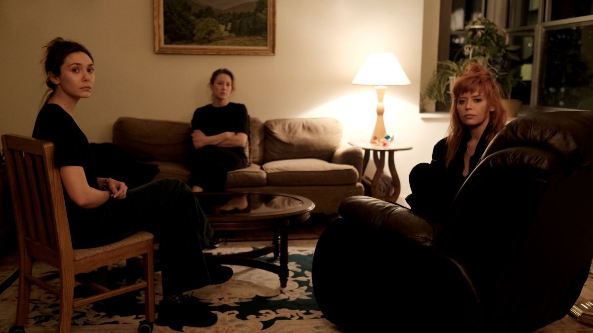 In a living room, Elizabeth Olsen as Christina sits on a chair on the left, Carrie Coon as Katie sits on the couch in the center, and Natasha Lyonne as Rachel sits on an armchair to the right in a scene from His Three Daughters