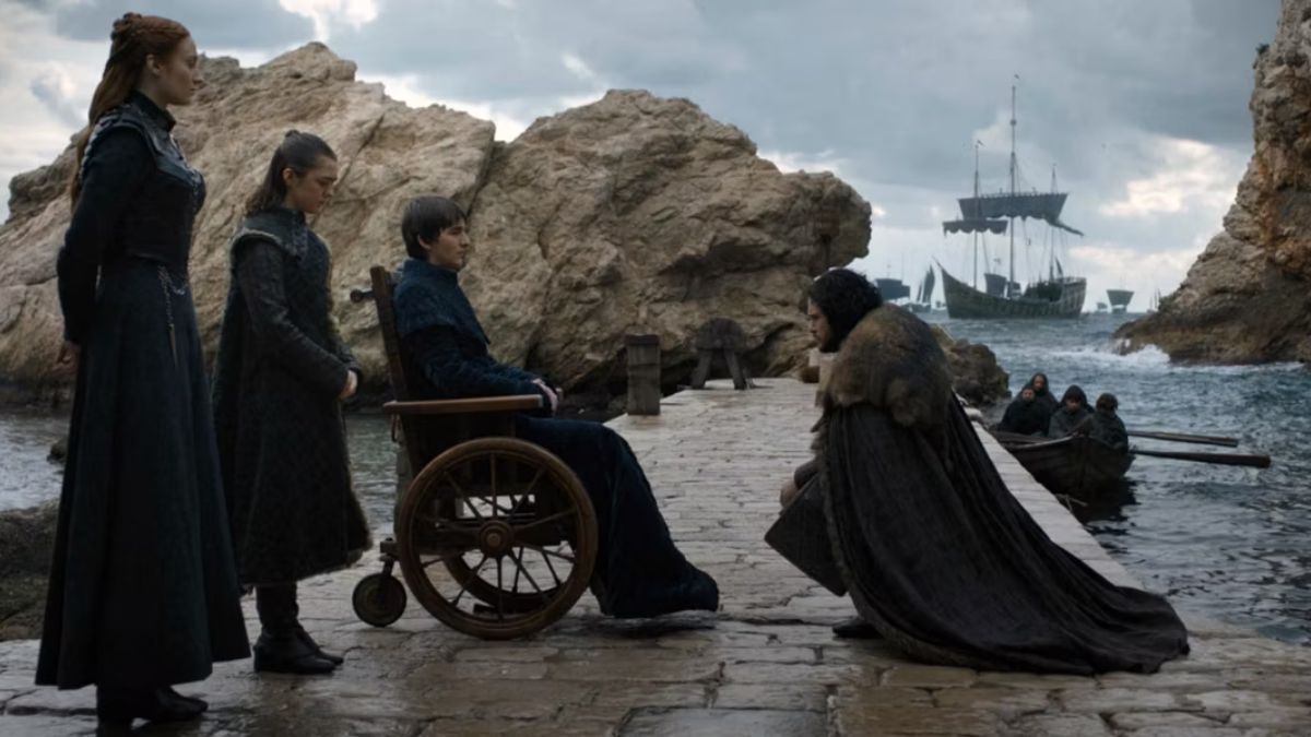 Sansa Stark and Arya Stark stand behind Bran Stark on his wheelchair as Jon Snow kneels to him on a deck by the sea.