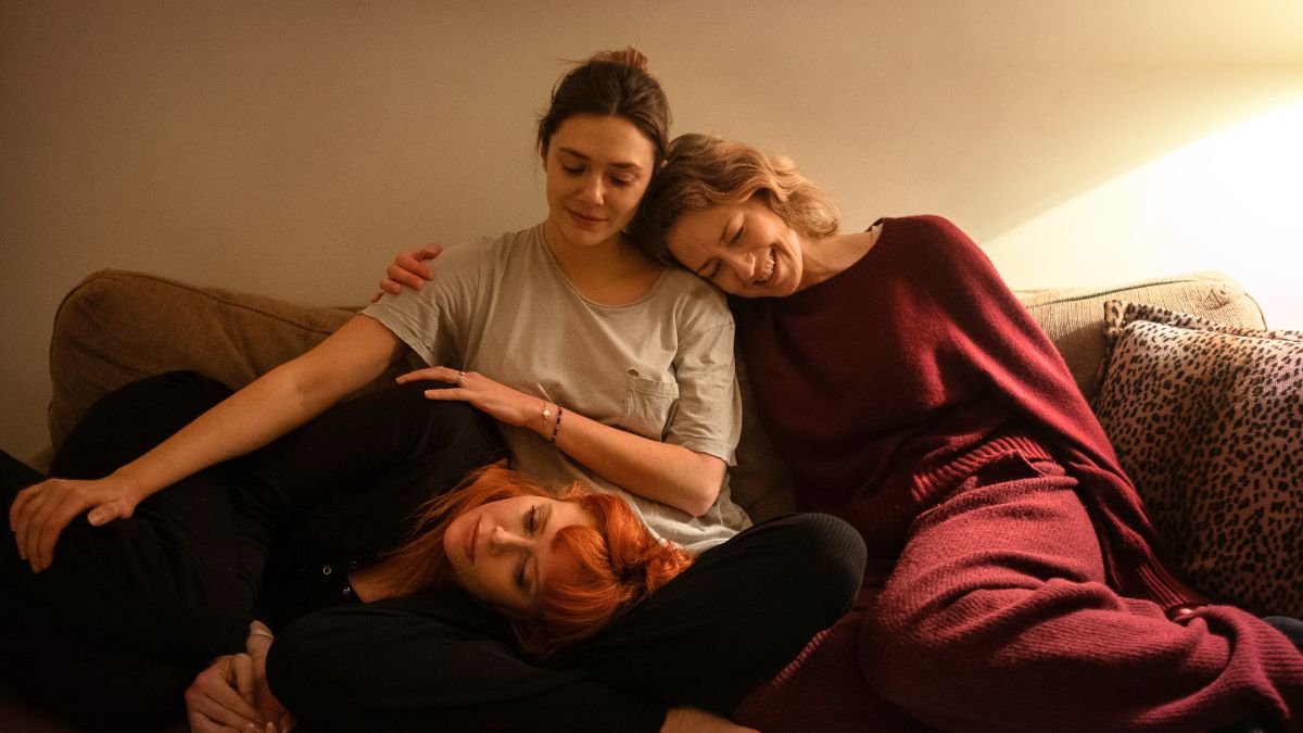 Elizabeth Olsen as Christina sits in the middle on a couch as Carrie Coon's Katie laughingly rests her head on her one shoulder, while Natasha Lyonne's Rachel rests her head in Olsen's lap in a scene from His Three Daughters