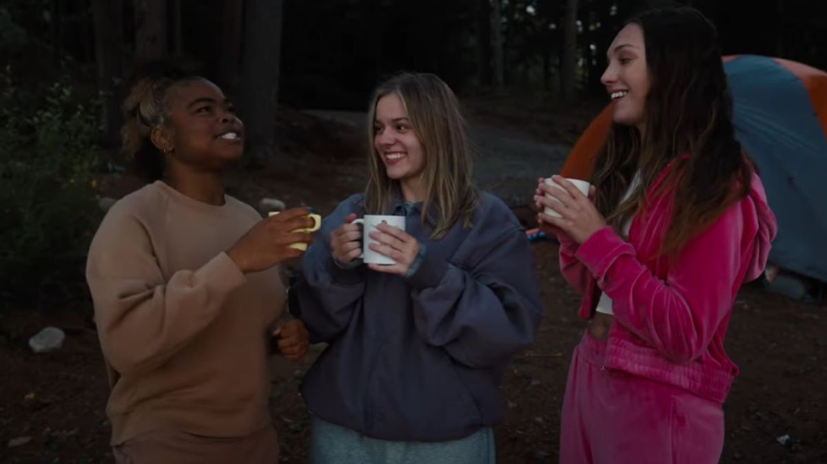 Image of Kerrice Brooks, Maisy Stella, and Maddie Ziegler in a scene from 'My Old Ass.' Brooks is a young, Black woman with dark hair streaked with blonde in a beige sweatshirt. Stella is a young white woman with shoulder-length blonde hair in a blue sweatshirt. Ziegler is a young white woman with long brown hair wearing a hot pink velvet track suit. They are all outside on a campsite holding mugs and smiling.