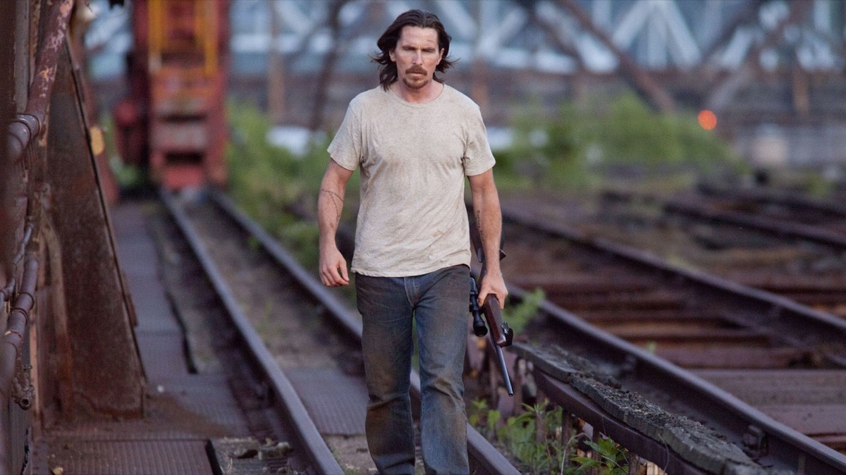 A blue collar man walks down train tracks with a scoped rifle in "Out of the Furnace"