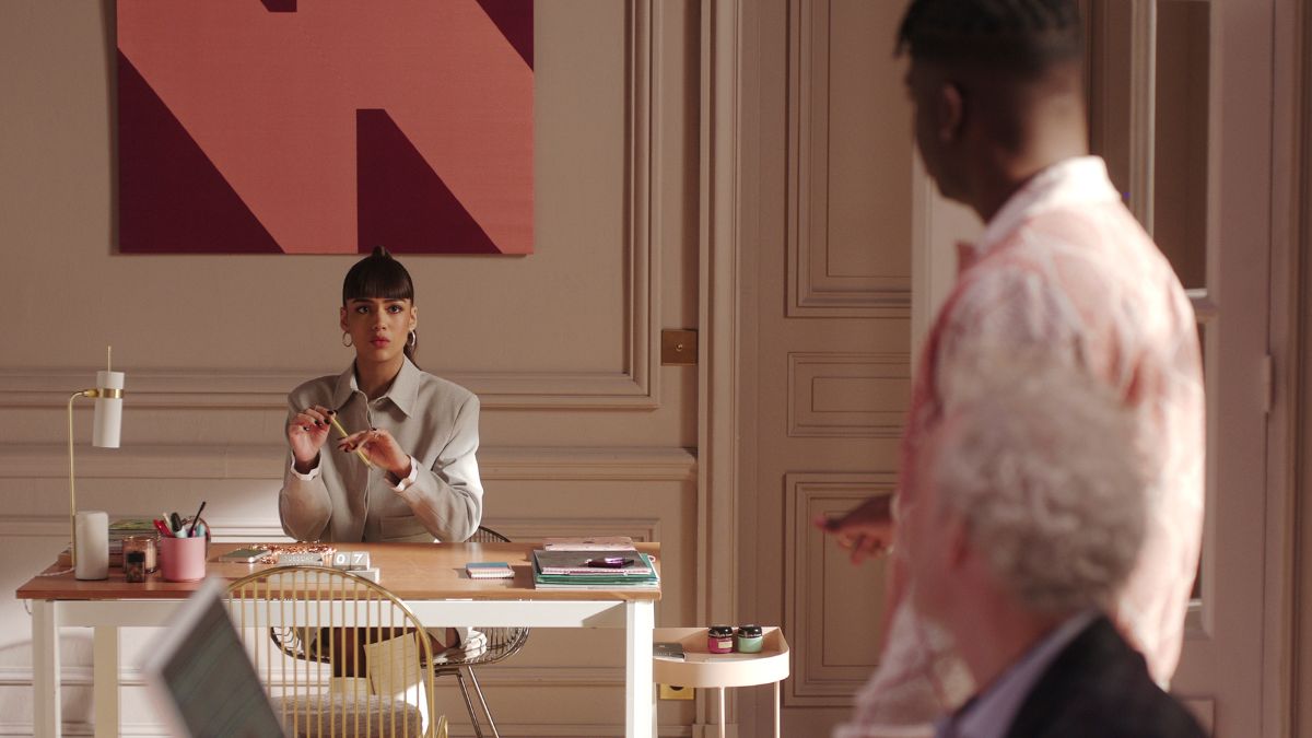 Thalia Besson as Genevieve sits at a desk as Samuel Arnold as Julien and Bruno Gouery as Luc look on in Emily In Paris season 4 part 2