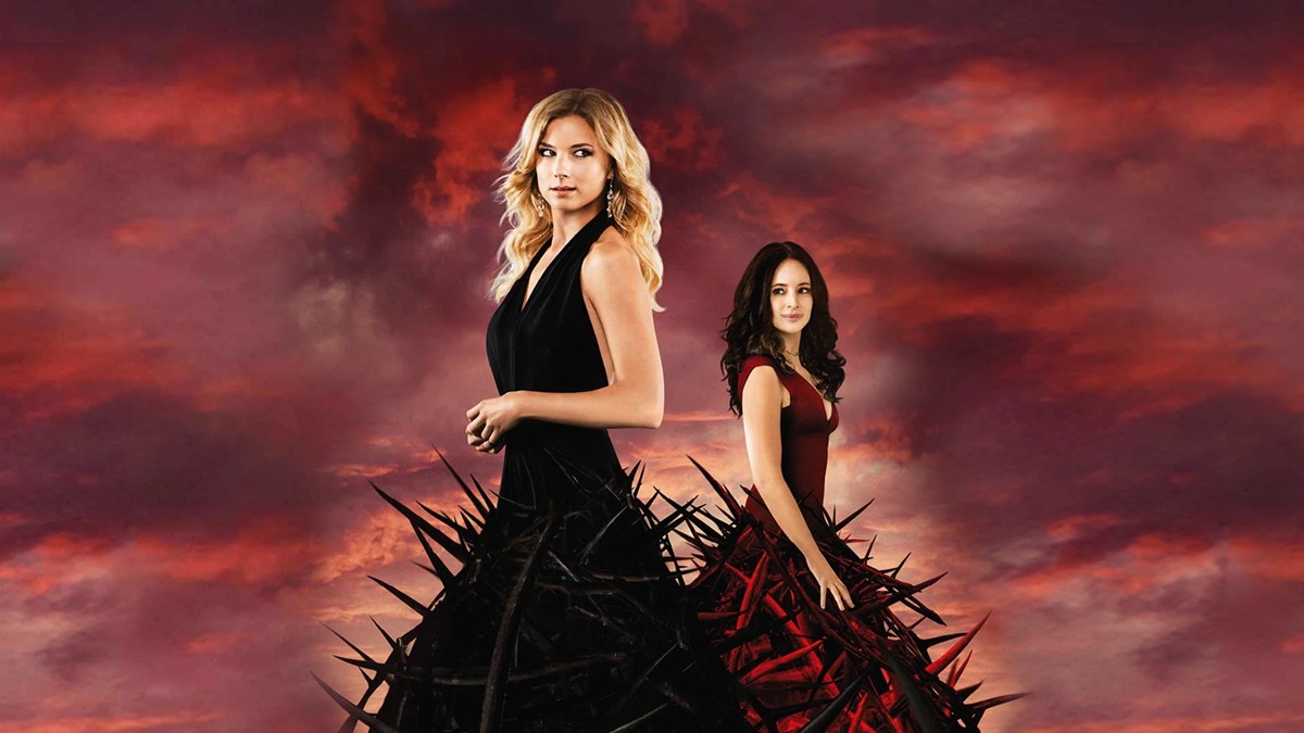 two women in fancy ball gowns standing against a deep red backdrop in 'Revenge' 