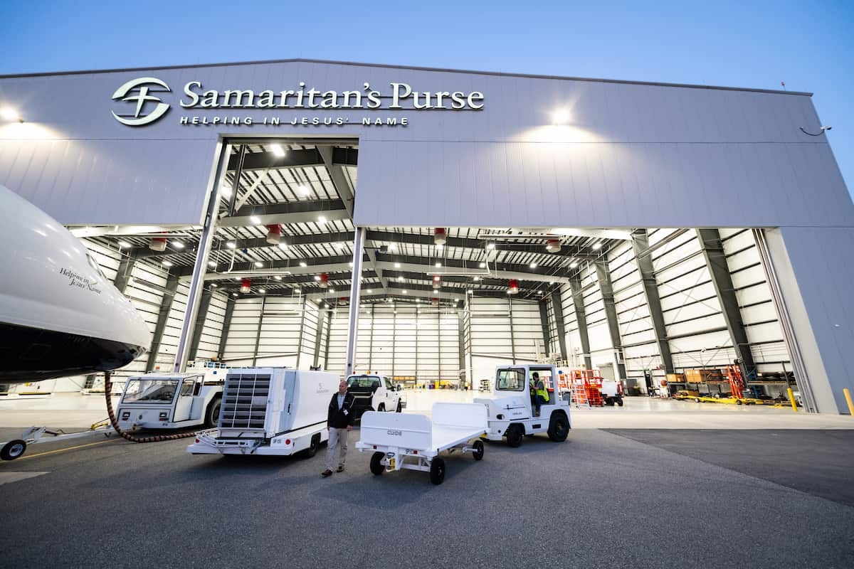 A Samaritan's Purse hangar is pictured, with "Helping in Jesus' Name" written on the building. Airport equipment and vehicles are parked in front, and the large open hangar is lit from the inside.