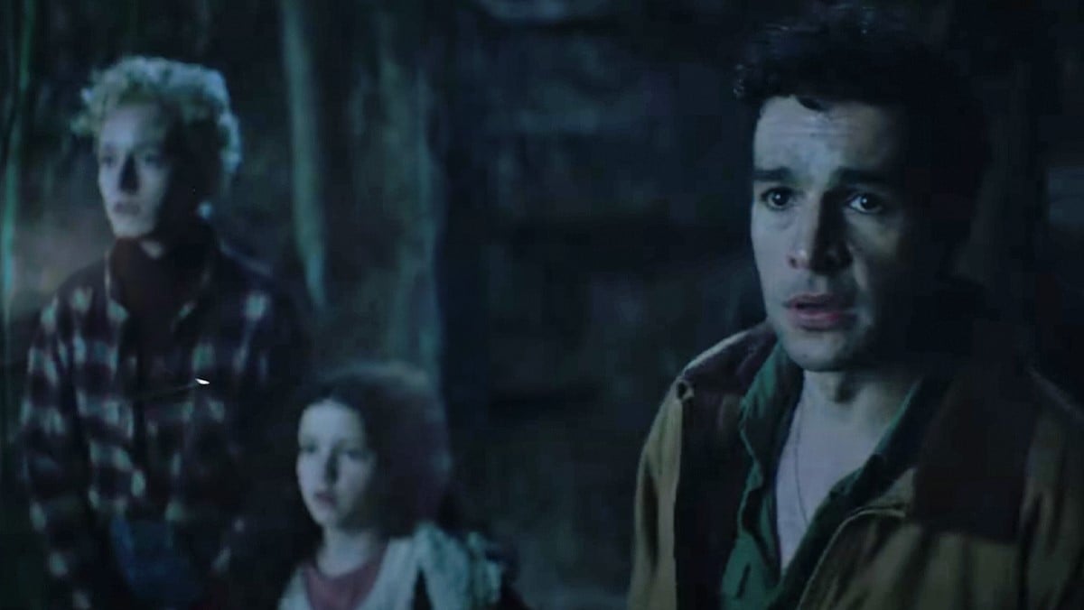 Julia Garner, Matilda Firth, and Christopher Abbott in a scene from the Universal film, 'Wolf Man.' They are all white. Garner is a woman with short, curly blond hair wearing a plaid button-down. Firth is a young girl with dark hair wearing a red t-shirt under a white sweater. Abbott is a man with short, dark hair wearing a green button-down under a brown jacket. They are standing in the woods.