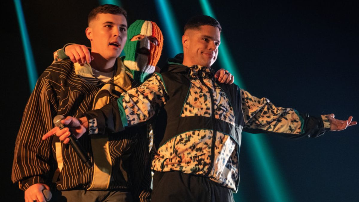 Three young men, one with a mask on his face, stand on a stage against flashing green lights