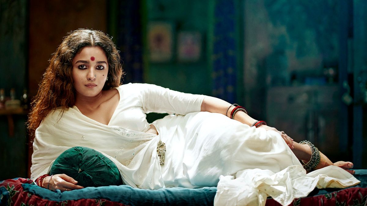 A woman clad in a white saree reclines on a bed, looking at the camera