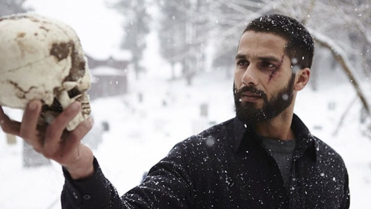 A man with a face injury stands in the snow, looking at a human skull in his hand