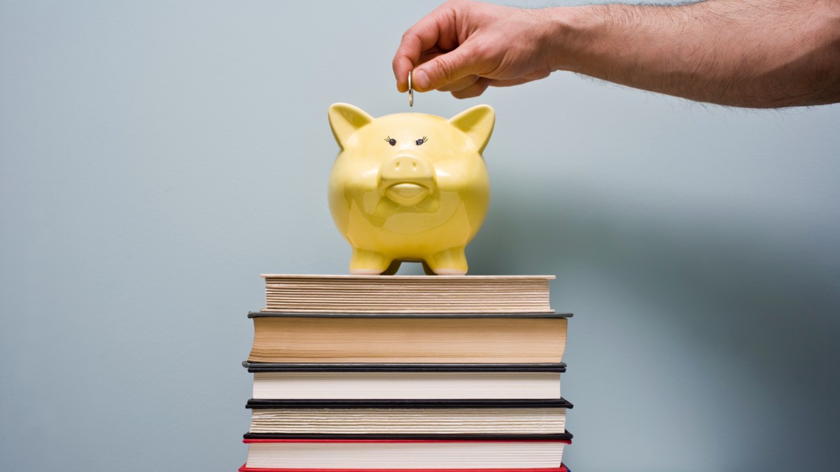 A hand puts coins into a piggy bank standing on top of a stack of books