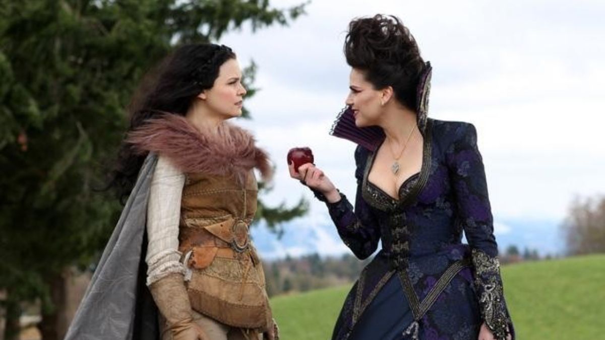 A woman dressed a in black gown offers another woman, dressed in brown furs, an apple.