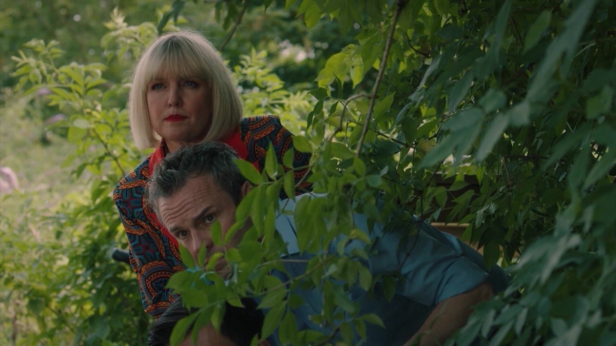 A middle-aged man and woman peer out from behind a bush in "Agatha Raisin"